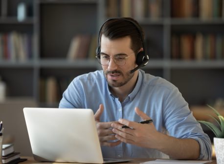 Confident man teacher coach wearing headset speaking, holding online lesson, focused student wearing glasses looking at laptop screen, studying, watching webinar training, listening to lecture